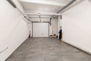 A clean, empty garage with a closed white door, grey tiled floor, and various tools and equipment against the walls. - Panoramic Sea-view Apartment in Le Conquet (Le Conquet)