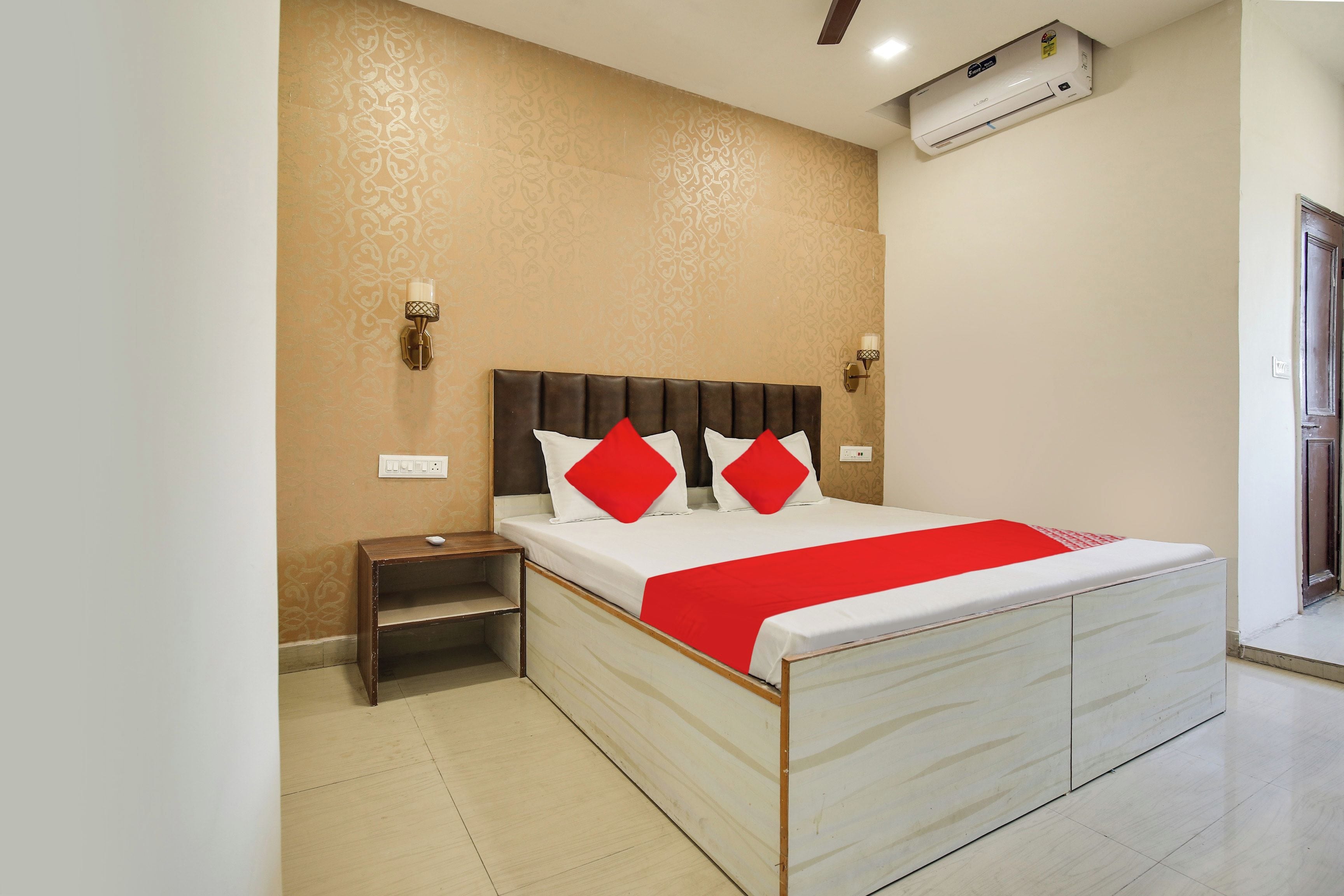 A hotel room with a bed, bedside table, nightstand, wall-mounted lamp, and an air conditioning unit.