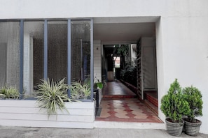 Entrada residencial com caminho de azulejos que leva à porta, plantas em vasos e janela com vista para a rua.