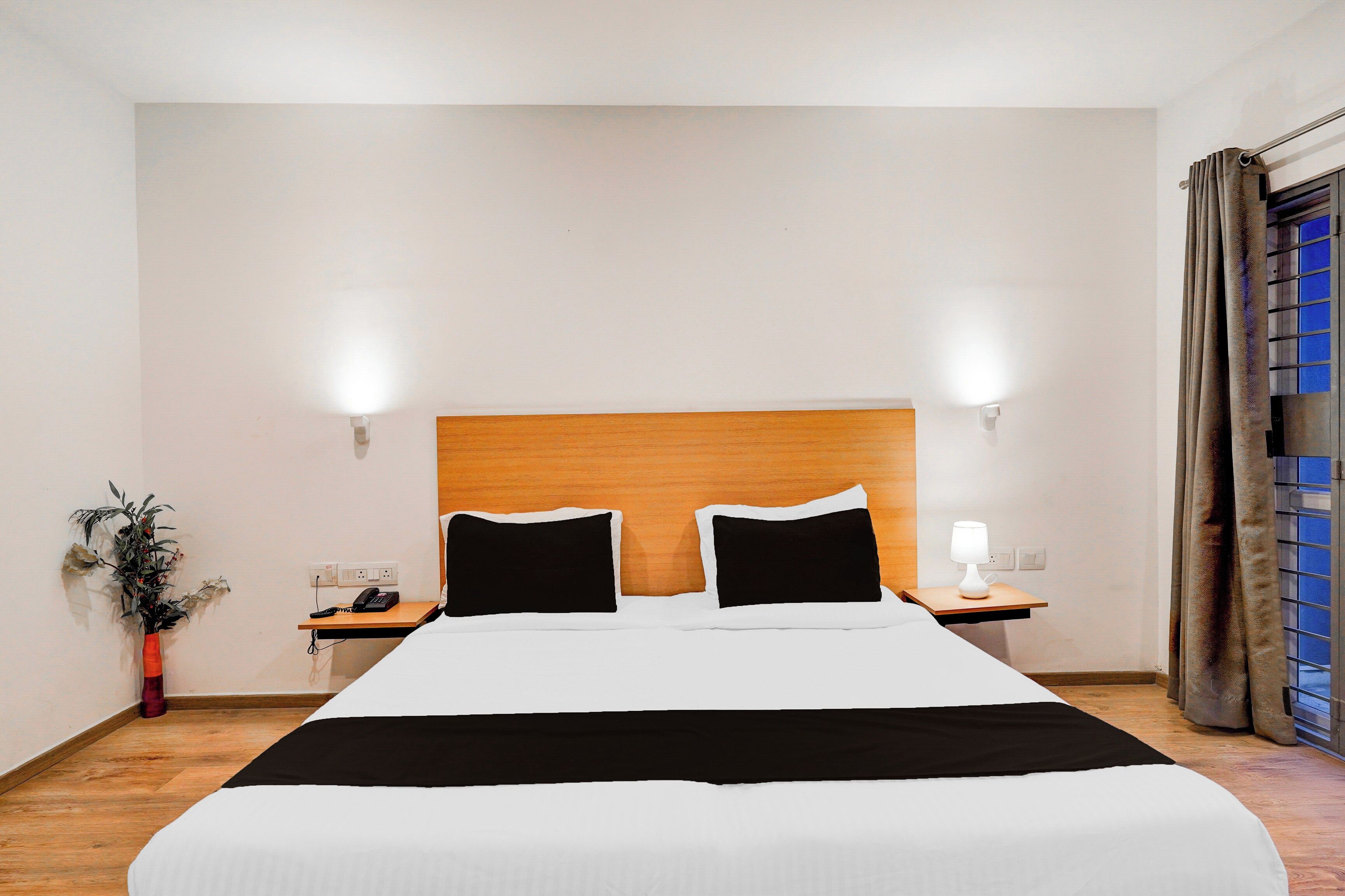 A hotel room with a large bed, bedside tables, a nightstand, a lamp, and a view of the outside through a window with curtains.