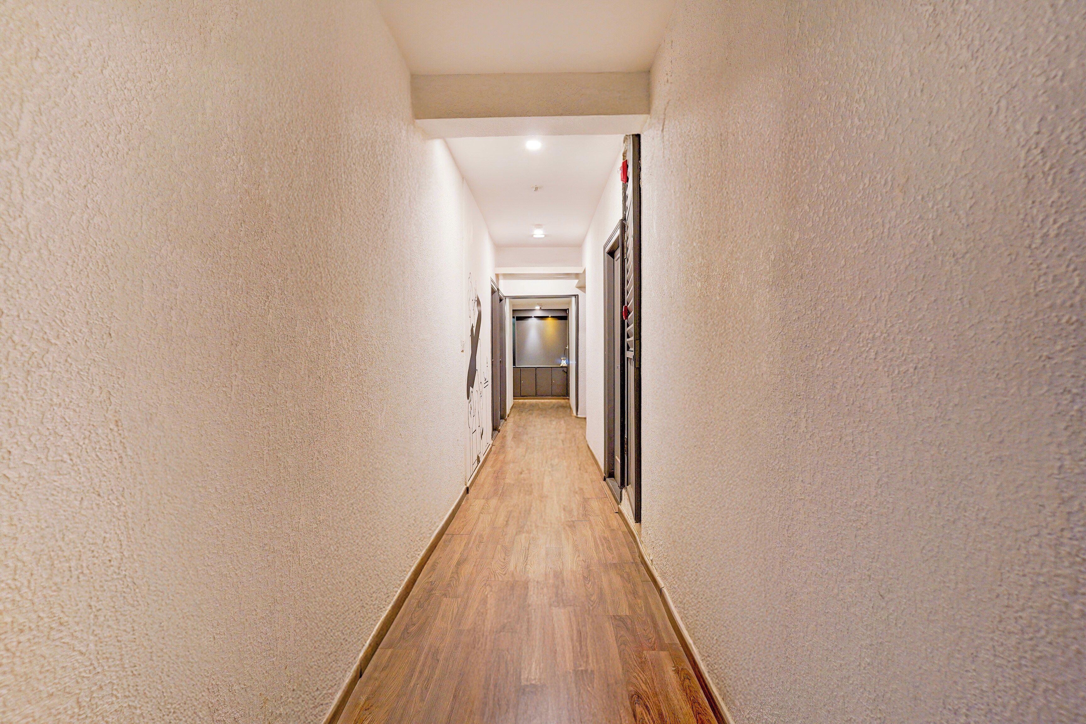 Un estrecho pasillo con suelo de madera y paredes blancas, que conduce a una habitación con una puerta y un ventanal.