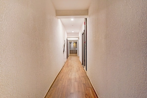 Um corredor estreito com piso de madeira e paredes brancas, dando acesso a um quarto com porta e janela.