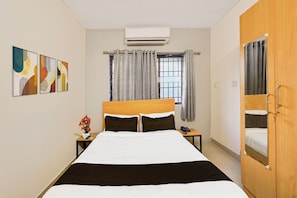 A hotel room with a bed, bedside table, a telephone, an air conditioning unit, a window with curtains, and a door. - Townhouse Nanganallur Near Chennai (Chennai)