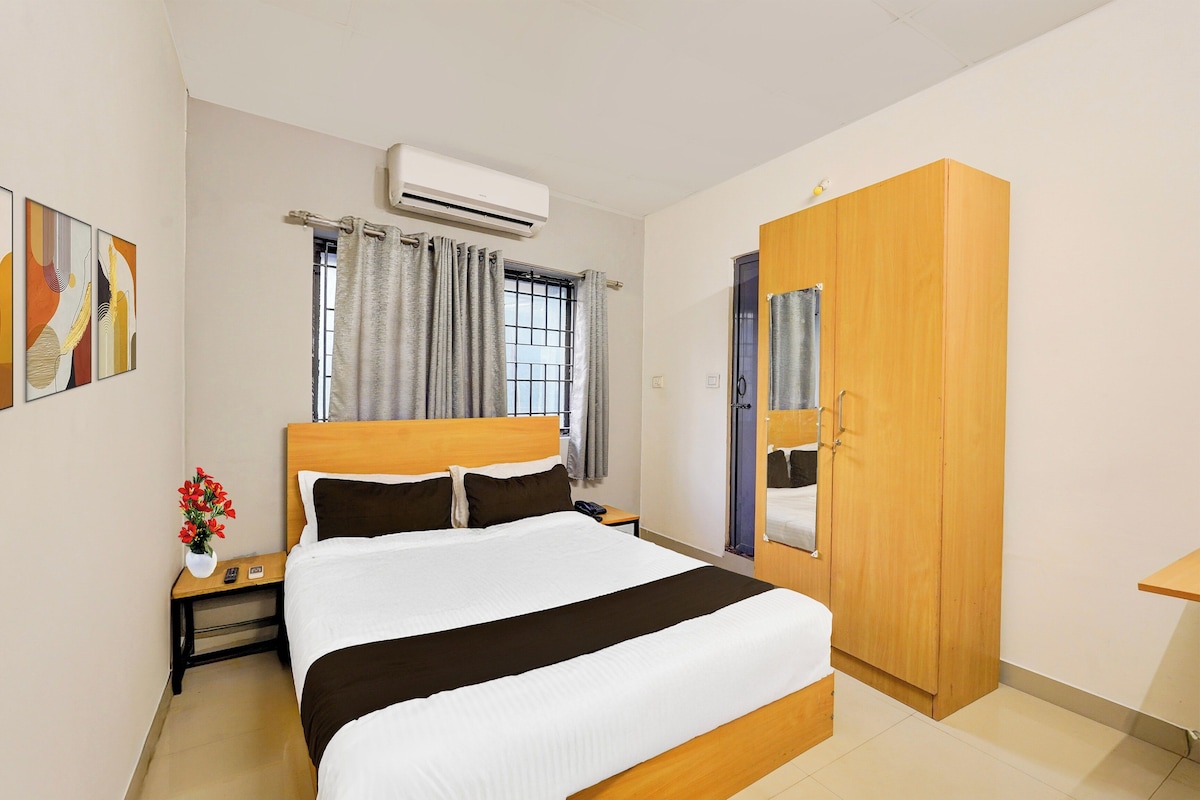 A hotel room with a bed, bedside table, nightstand, an air conditioning unit, a wardrobe, and a window with curtains.