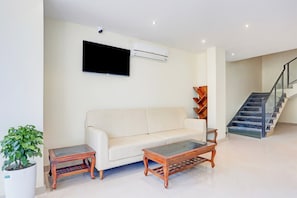 A living room with a sofa, two wooden side tables, a potted plant, a mounted flat-screen TV, and a staircase. - Townhouse ISBT Dehradun (Dehradun)