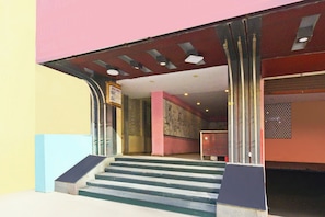 Entrada de um edifício com um conjunto de degraus que levam a uma porta, uma placa acima da porta e uma parede com um desenho padronizado.