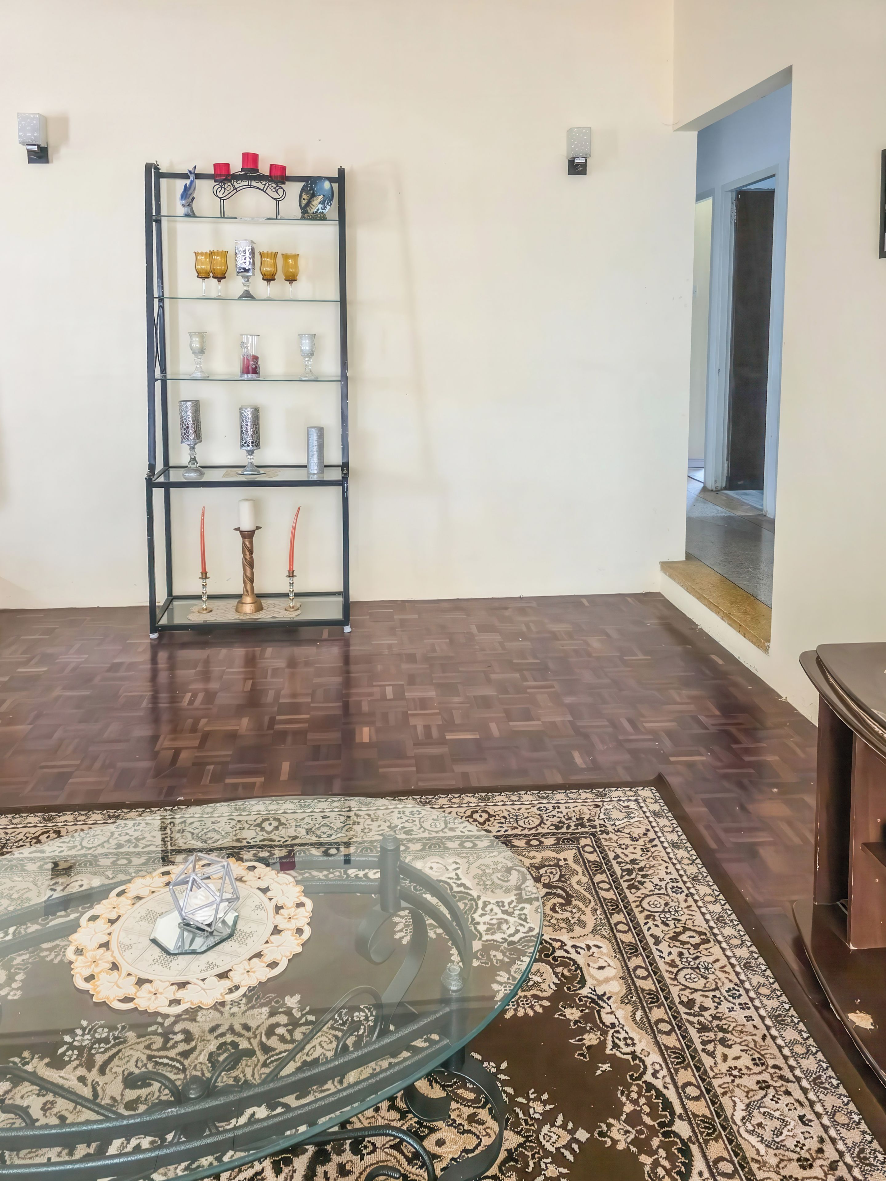A room with a glass-top table, a patterned rug, a metal shelf with various items, and a wooden floor.