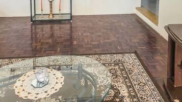 A room with a glass-top table, a patterned rug, a metal shelf with various items, and a wooden floor.