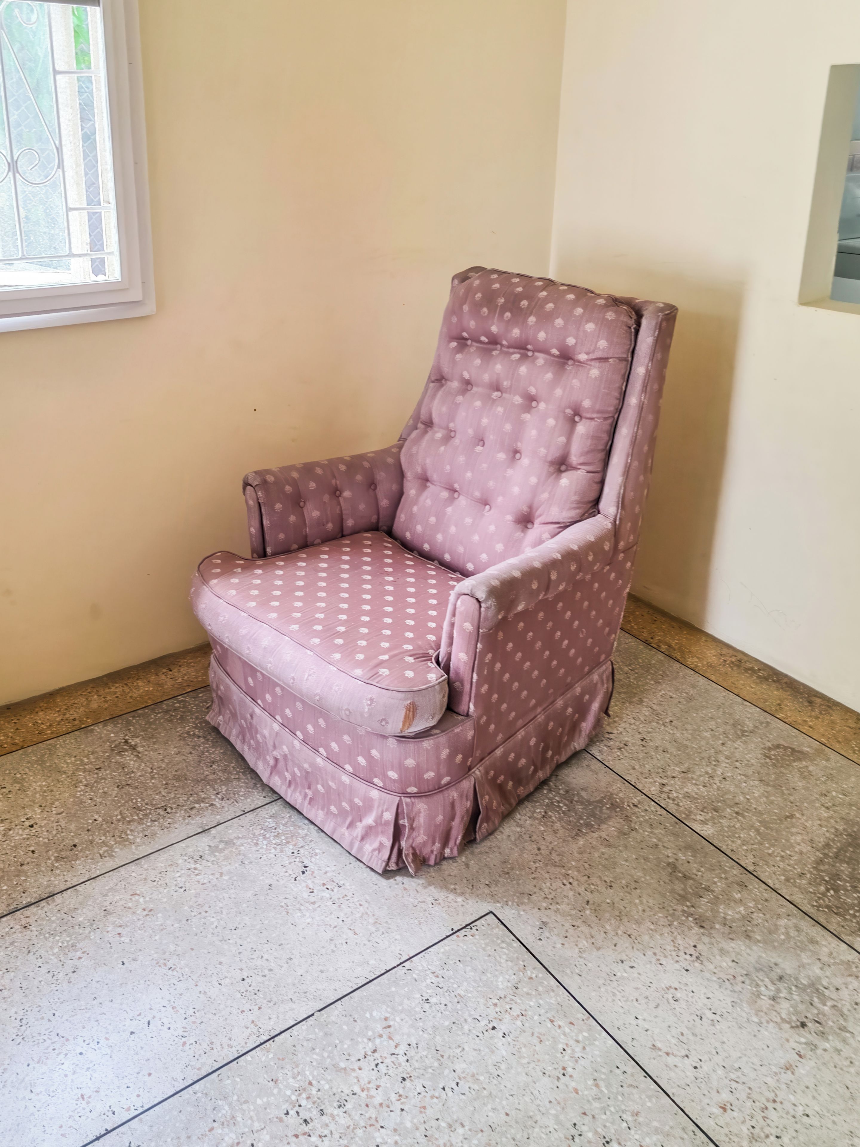 A corner room with a single cushioned chair, a window with a decorative grille, and a plain wall.