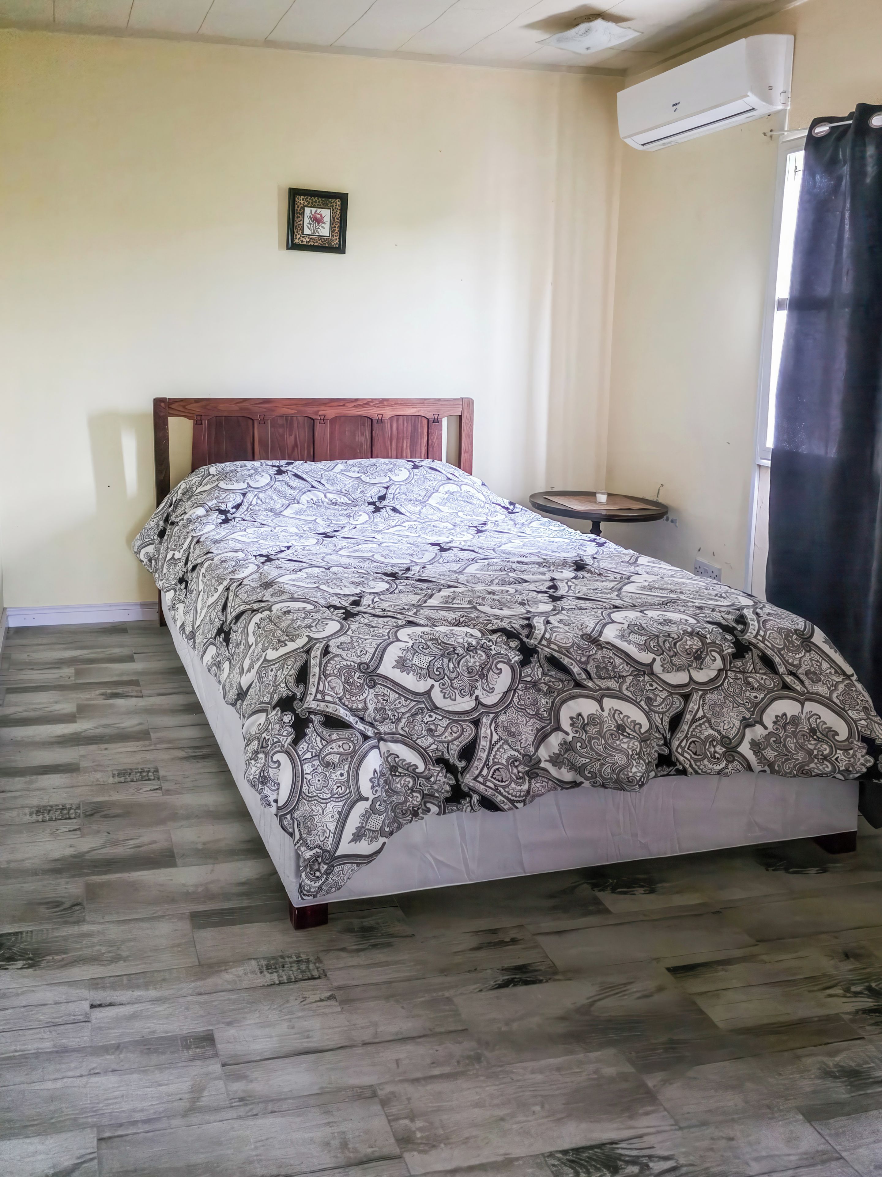 A bedroom with a wooden bed, patterned bedspread, a small round table, and an air conditioning unit.