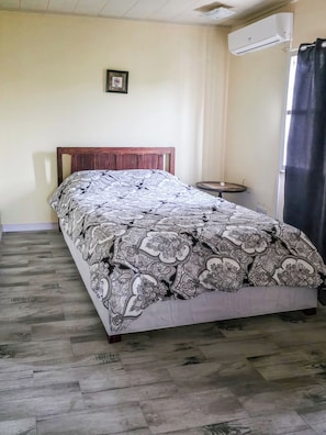 A bedroom with a wooden bed, patterned bedspread, a small round table, and an air conditioning unit. - Home Away From Home (Arnos Vale)