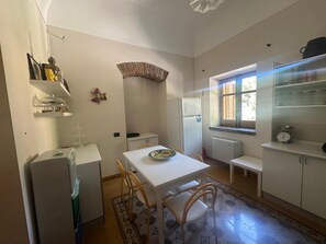 Cozinha compacta com mesa branca, cadeiras, geladeira e janela com persianas.