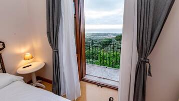 A hotel room with a bed, a small table with a lamp, and a view of a balcony with greenery and a distant cityscape.