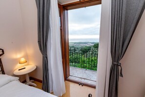 Une chambre d’hôtel avec un lit, une petite table avec une lampe et une vue sur un balcon agrémenté de verdure et sur le paysage urbain au loin.