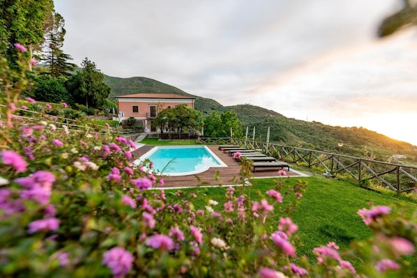 A pool area with a wooden deck and lounge chairs, surrounded by a fence and lush greenery.