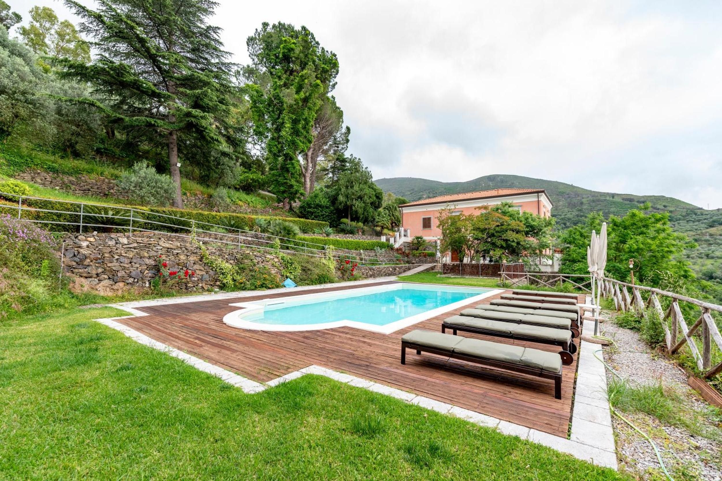 Una piscina con cubierta de madera y sillas de descanso, rodeada por un muro de piedra y abundante vegetación.