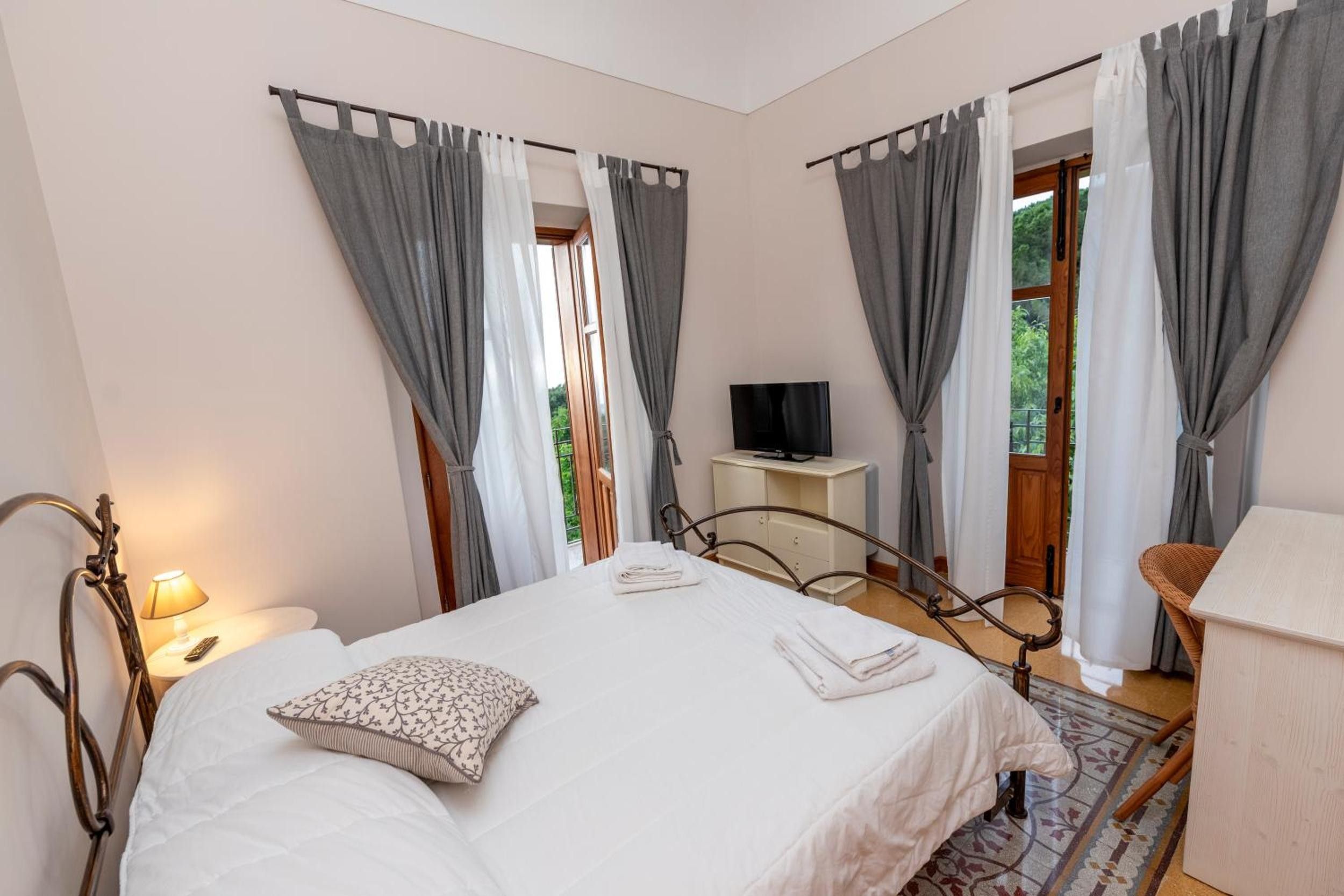 Une chambre à coucher avec un lit, une table de chevet avec une lampe, un téléviseur posé sur un meuble et une fenêtre avec des rideaux.