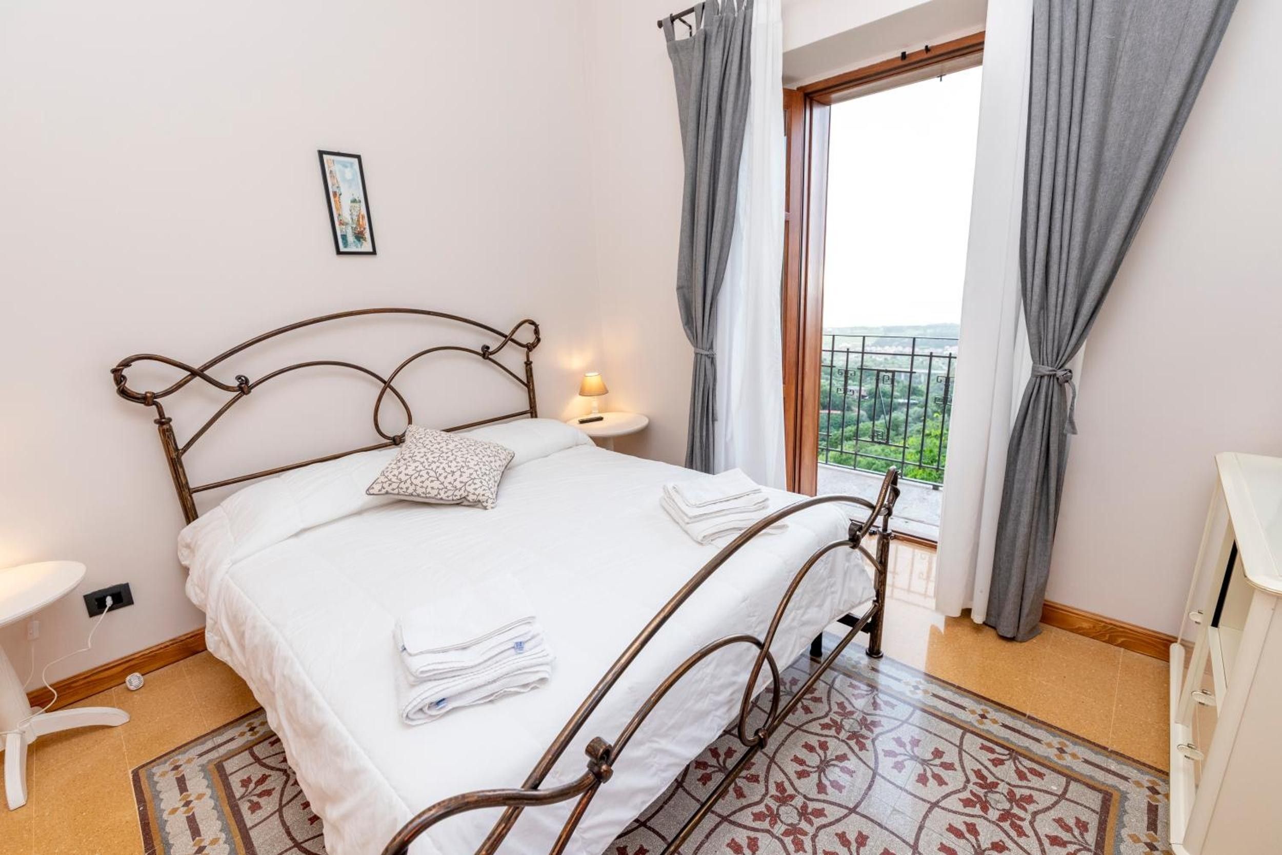 A bedroom with a metal bed frame, a bedside table with a lamp, a window with curtains, and a view of greenery outside.