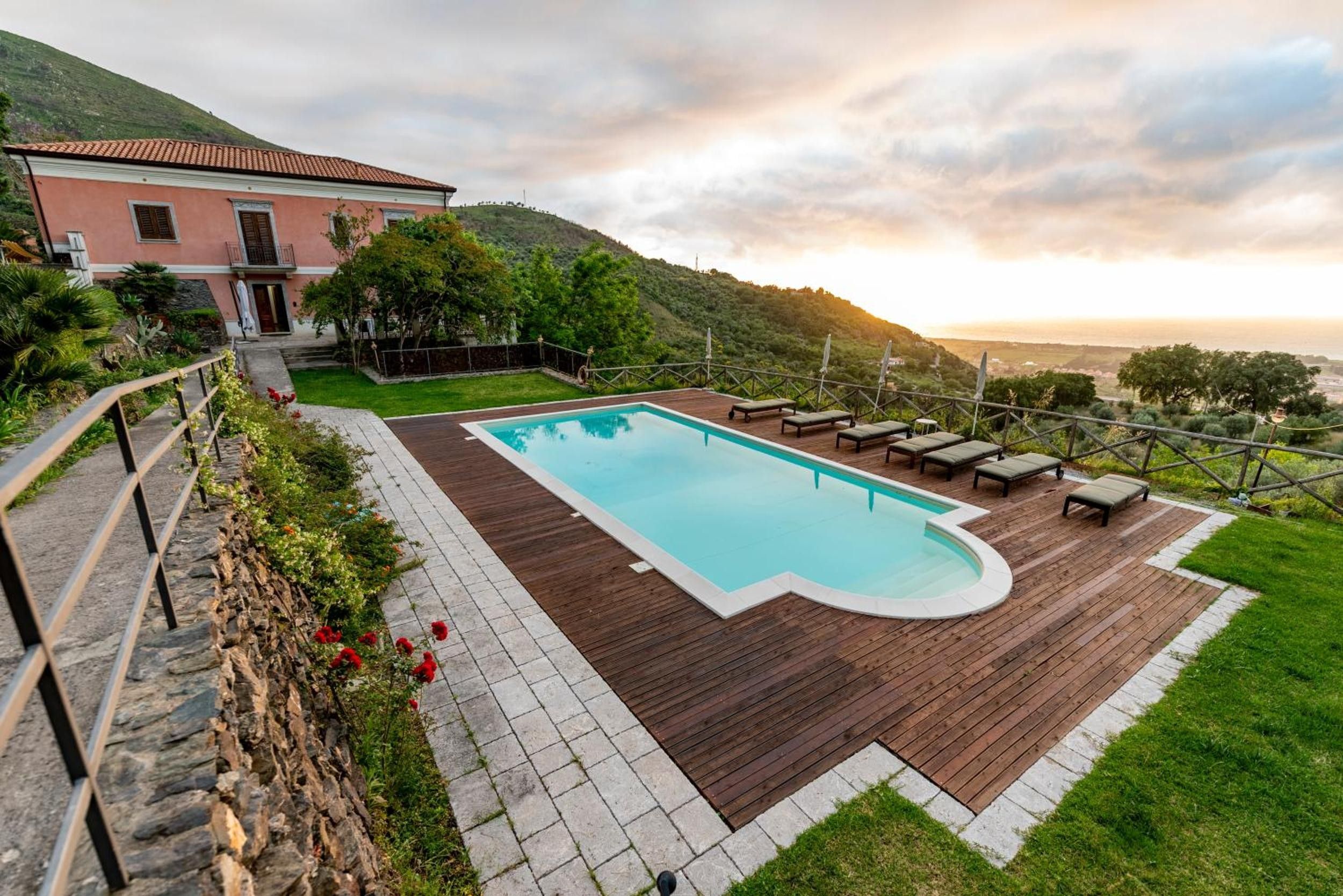 A swimming pool with a wooden deck, surrounded by a stone wall and a grassy area.