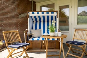 A wooden deck with a striped chair and two wooden chairs, a small table with a white vase and a plant, and a brick wall. - Holiday apartment with 2 bedrooms (Westerland - Sylt)