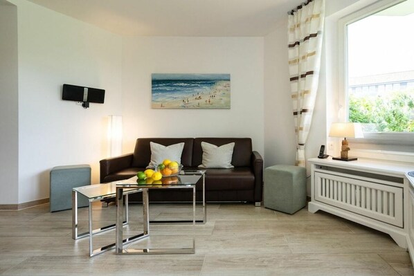 Uma sala de estar moderna com um sofá marrom, uma mesa de centro de vidro com frutas, um banquinho cinza, uma pintura de uma paisagem praiana e uma janela com cortinas.