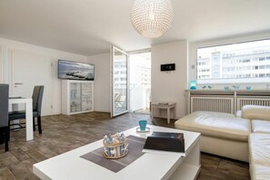 A modern living room with a white sofa, a dining table with chairs, a coffee table with decorative items, a large window with a view of buildings, and a chandelier. - Sylt Residence Apartment 5 (Sylt Ortsteil Westerland)