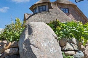 Uma pedra com a inscrição 'York's Hü', um edifício com telhado de palha e um muro de pedra.
