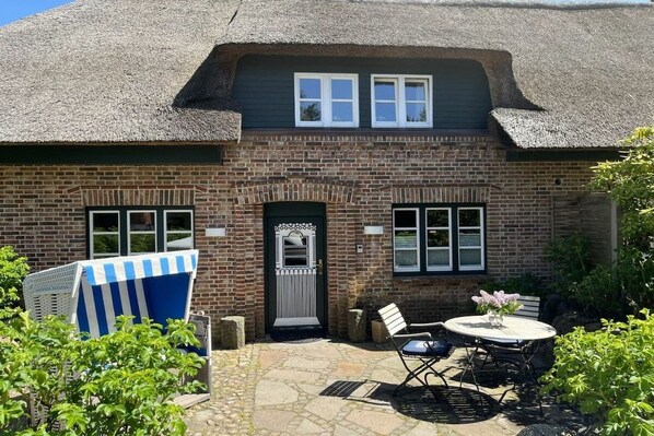 A brick house with a thatched roof, a front door, and a small patio with a table and chairs. - Holiday apartment in Sylt with 1 bedroom (Sylt \/ Alt Westerland)
