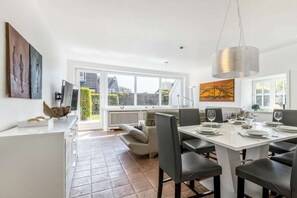 A modern dining area with a white table set for four, a large window with a view of the outside, and a hanging light fixture. - Holiday home in Sylt with swimming pool (Sylt)