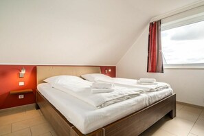 A bedroom with a wooden bed, white bedding, a nightstand with a lamp, and a window with curtains. - Lontargos upper floor apartment (Göhren-Lebbin)