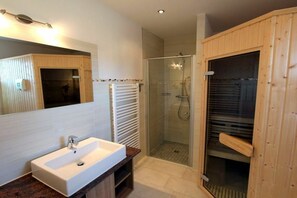 Ein modernes Badezimmer mit einem weißen Waschbecken, einer Glasdusche und einer Holzsauna.