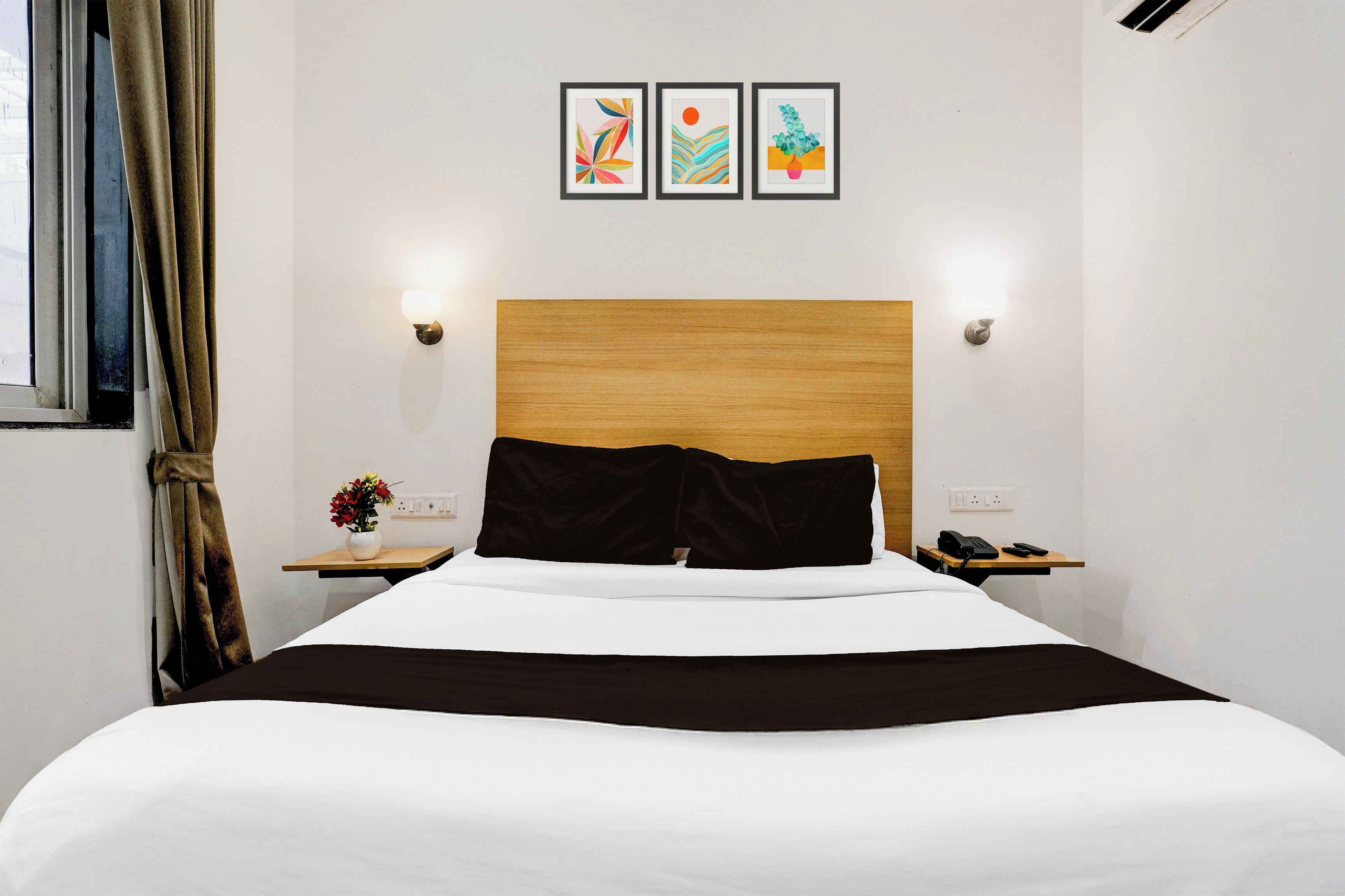 Une chambre d’hôtel avec un lit, une table de chevet avec un téléphone, un vase de fleurs et trois œuvres d’art encadrées accrochées au mur.
