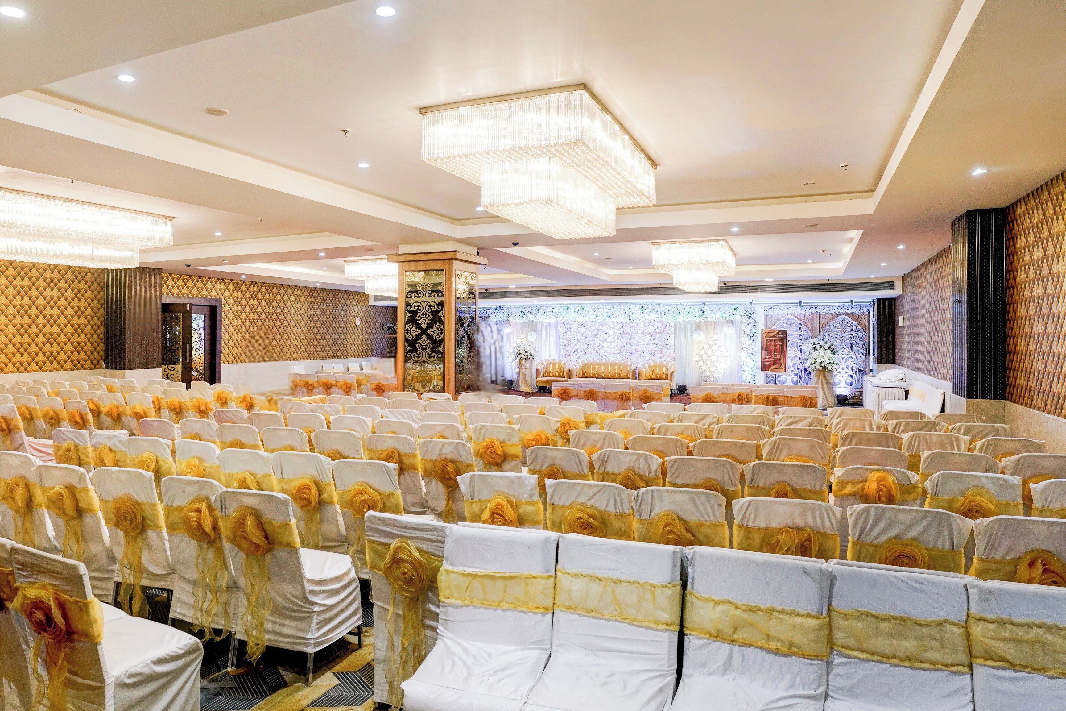 Un salón de banquetes con filas de sillas, un escenario con decoraciones florales y una araña de cristal.