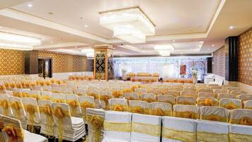 Un salón de banquetes con filas de sillas, un escenario con decoraciones florales y una araña de cristal.