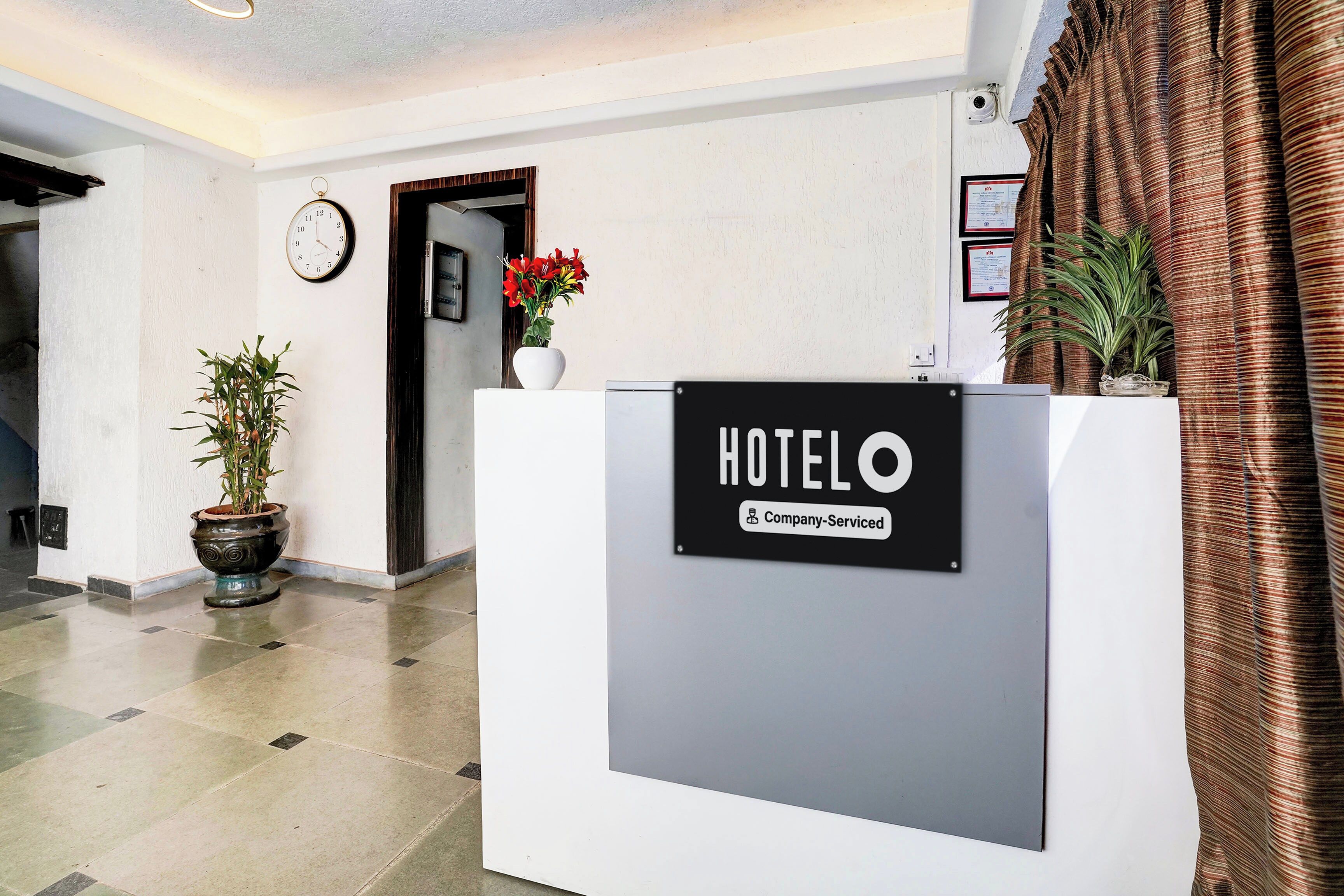 A hotel reception desk with a sign indicating it is company-serviced. There is a clock on the wall and a potted plant to the left.