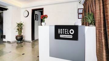 A hotel reception desk with a sign indicating it is company-serviced. There is a clock on the wall and a potted plant to the left.