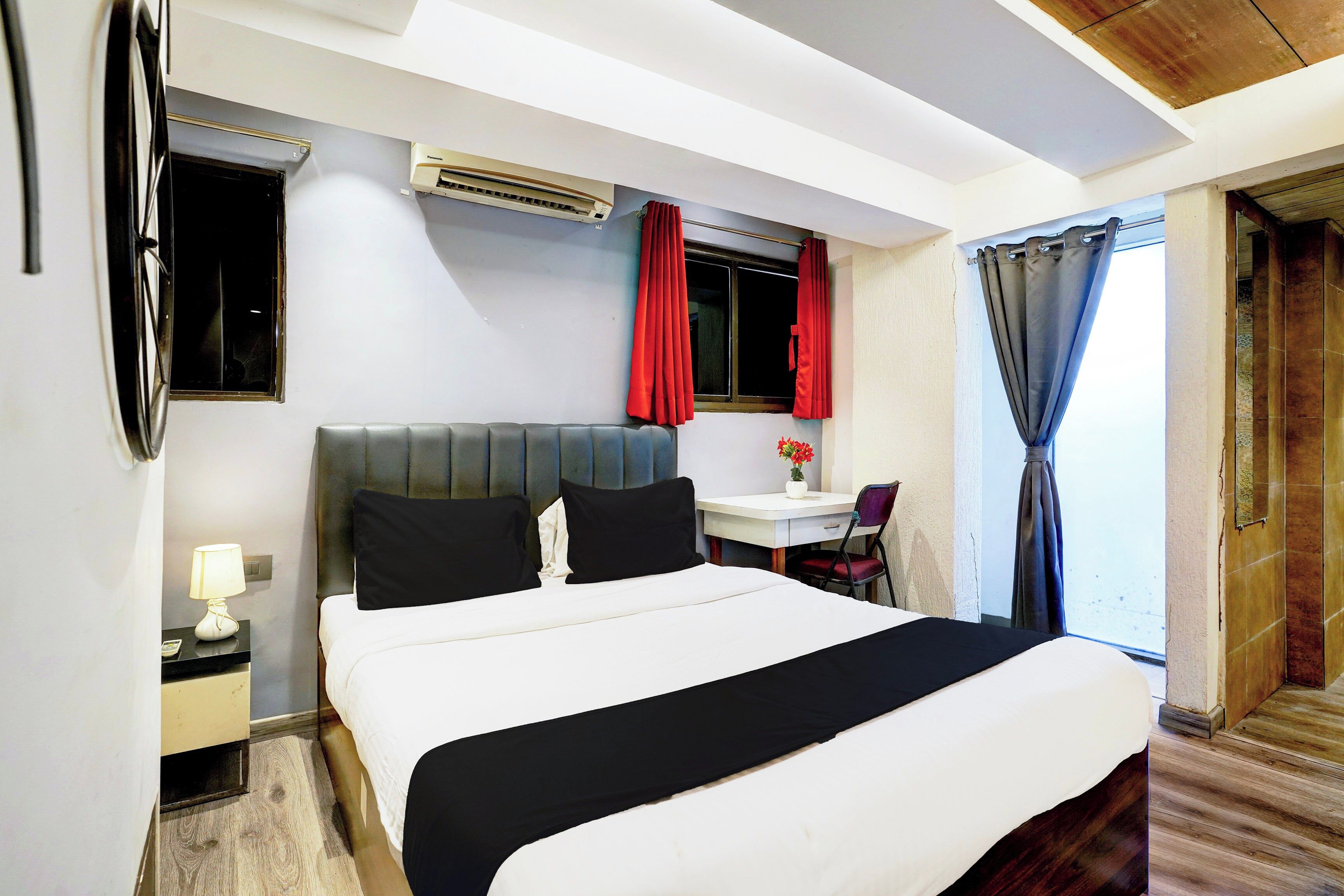 A hotel room with a bed, a desk, a chair, a lamp, a window with red curtains, and an air conditioning unit.