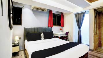 A hotel room with a bed, a desk, a chair, a lamp, a window with red curtains, and an air conditioning unit.