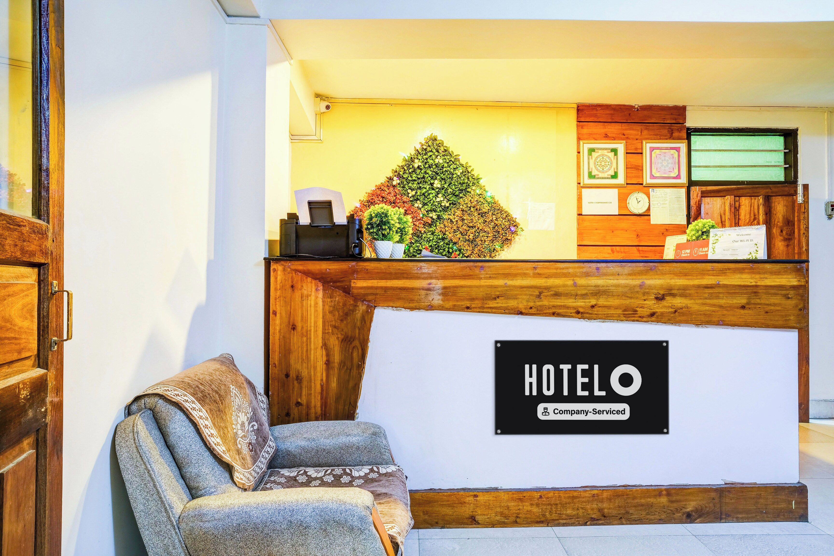 Un comptoir d’accueil d’hôtel avec une enseigne indiquant « HÔTEL O » et une étiquette « Service aux entreprises ». On y trouve un comptoir, une chaise et une plante décorative.