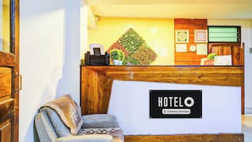 Un comptoir d’accueil d’hôtel avec une enseigne indiquant « HÔTEL O » et une étiquette « Service aux entreprises ». On y trouve un comptoir, une chaise et une plante décorative.