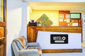 Mostrador de recepción de hotel con un letrero que dice 'HOTEL O' y una etiqueta que indica 'Servicio de empresa'. Se observa un mostrador, una silla y una planta decorativa.