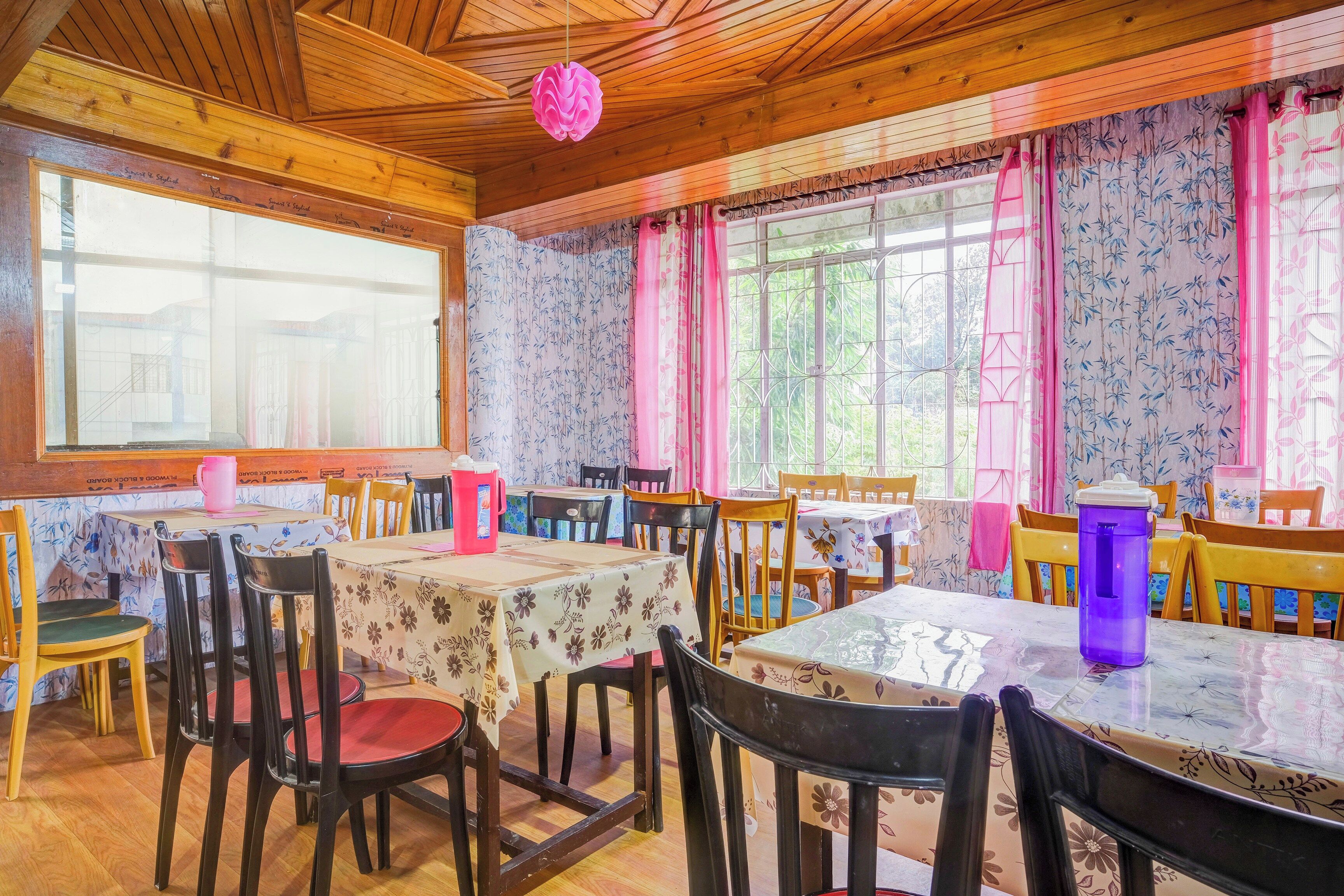Un comedor con mesas y sillas de madera, amplias ventanas con cortinas florales y techo de madera.