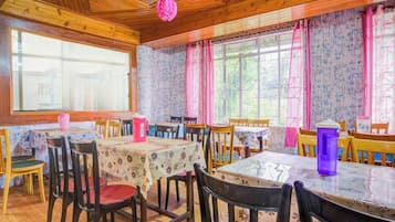 Un comedor con mesas y sillas de madera, amplias ventanas con cortinas florales y techo de madera.
