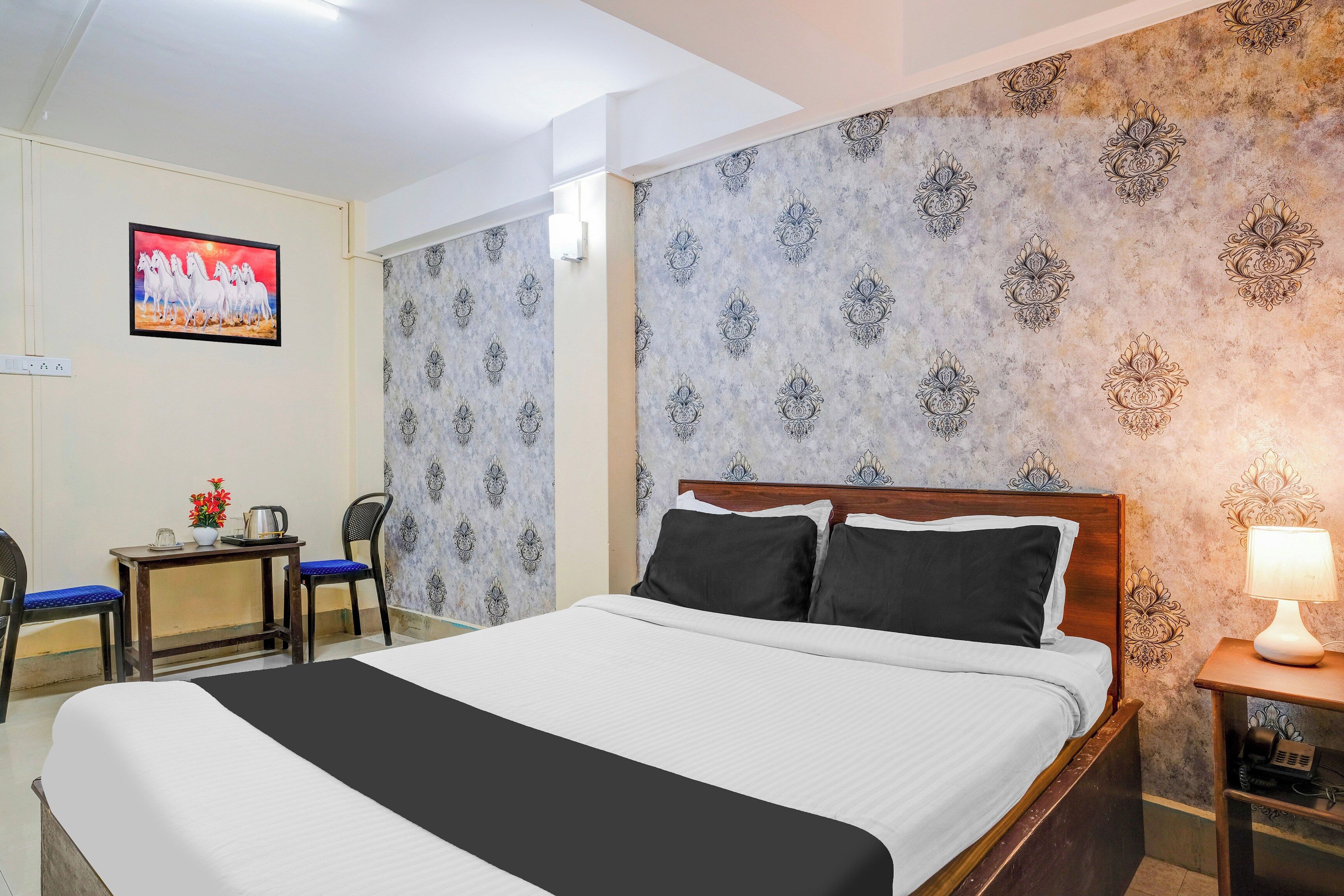 Ein Hotelzimmer mit einem Bett, einem Schreibtisch mit Stuhl, einem an der Wand befestigten Fernseher und ornamental gestalteter Tapete.