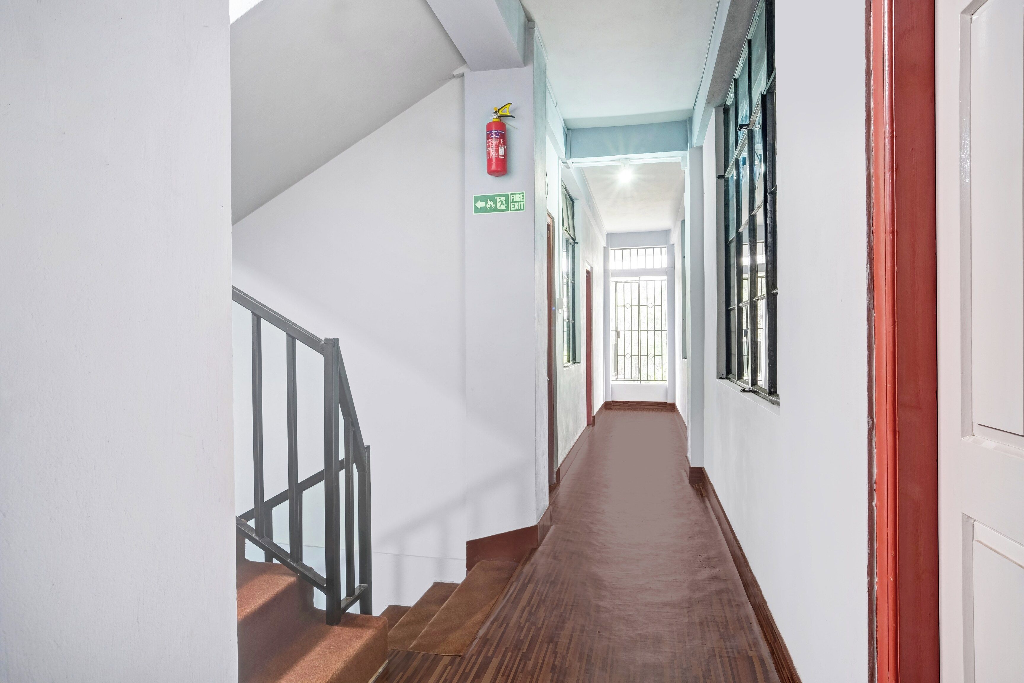 Un couloir avec un escalier en bois, un panneau indiquant la sortie de secours et une fenêtre avec des barreaux en métal.