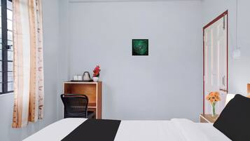 A hotel room with a bed, a chair, a desk with a kettle, a small plant, and a wall decoration.