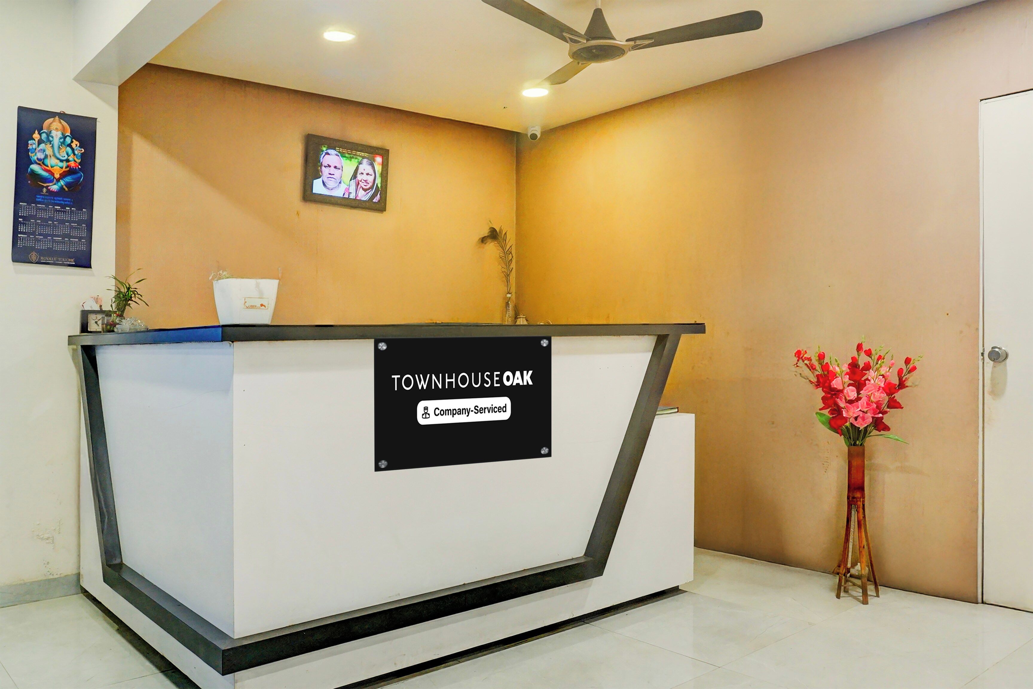 A reception desk with a sign indicating it is company-serviced. There is a ceiling fan above and a framed picture on the wall.