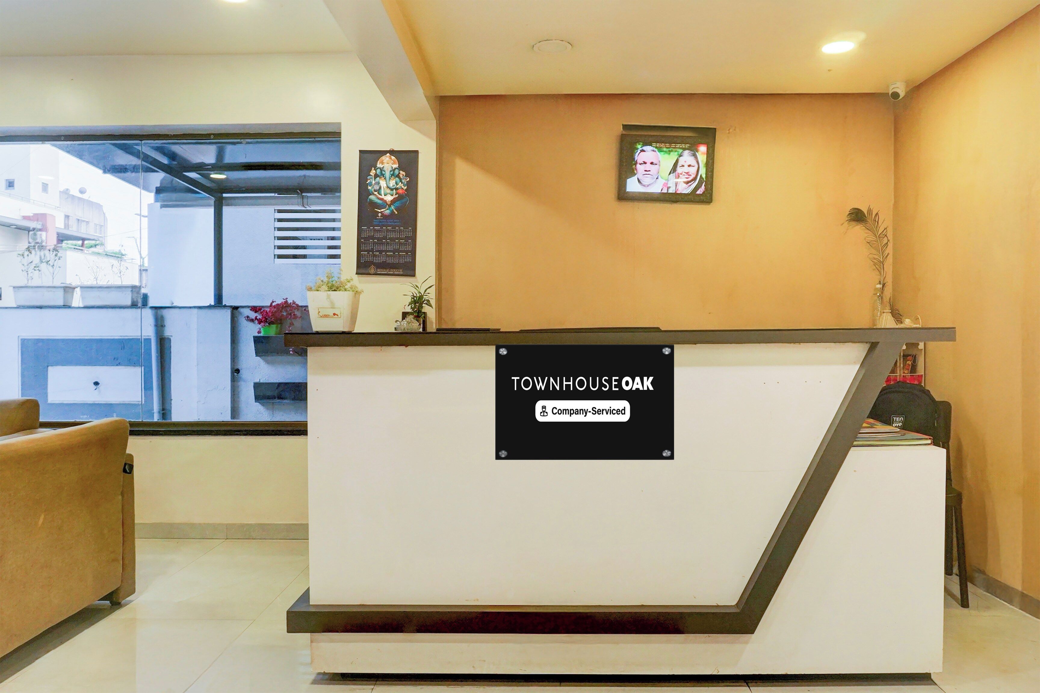 A reception desk with a sign indicating it is company-serviced. There are potted plants and a framed picture on the wall.