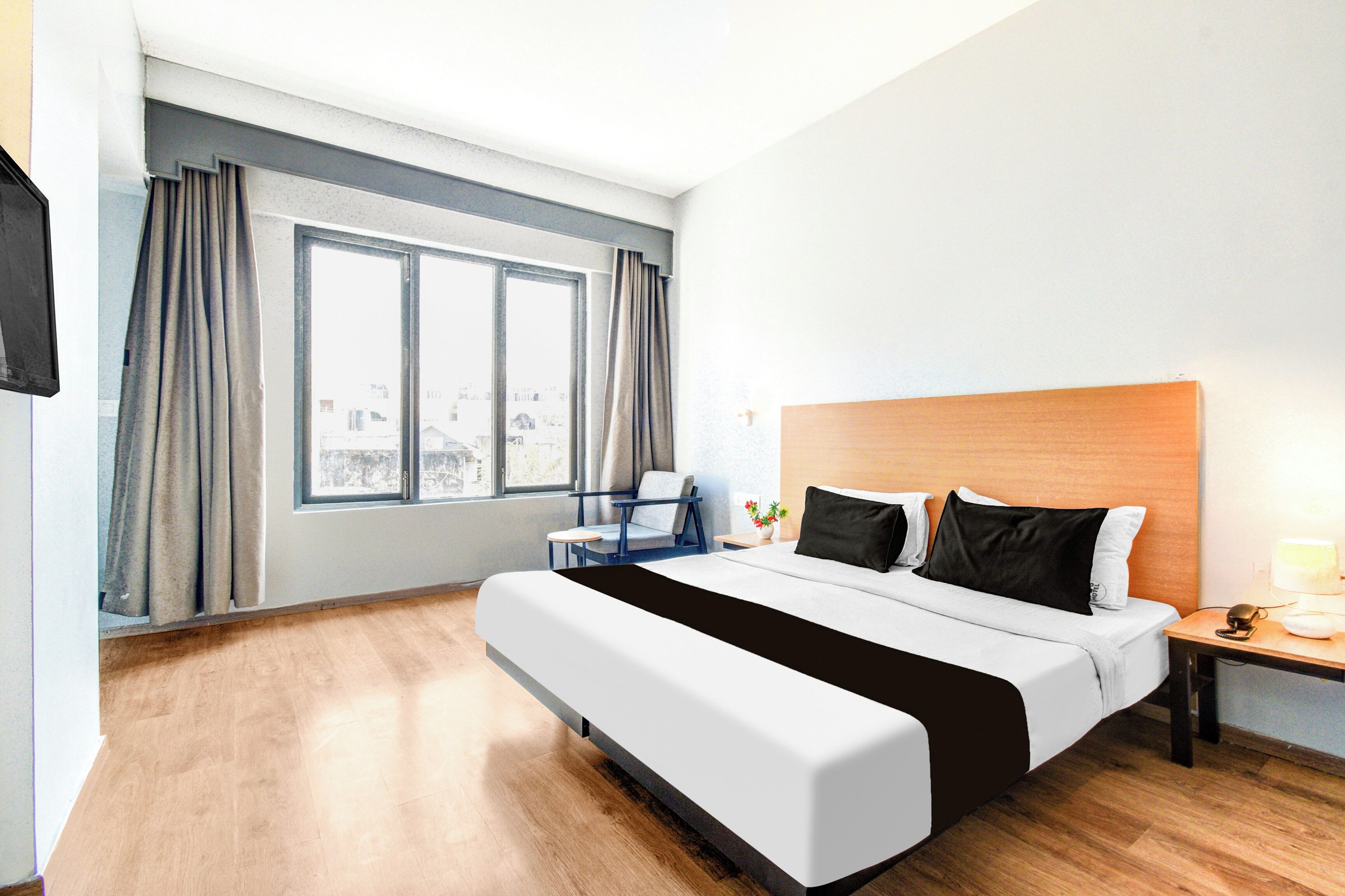 A hotel room with a large bed, a nightstand, a chair, a window with curtains, and a view of buildings outside.