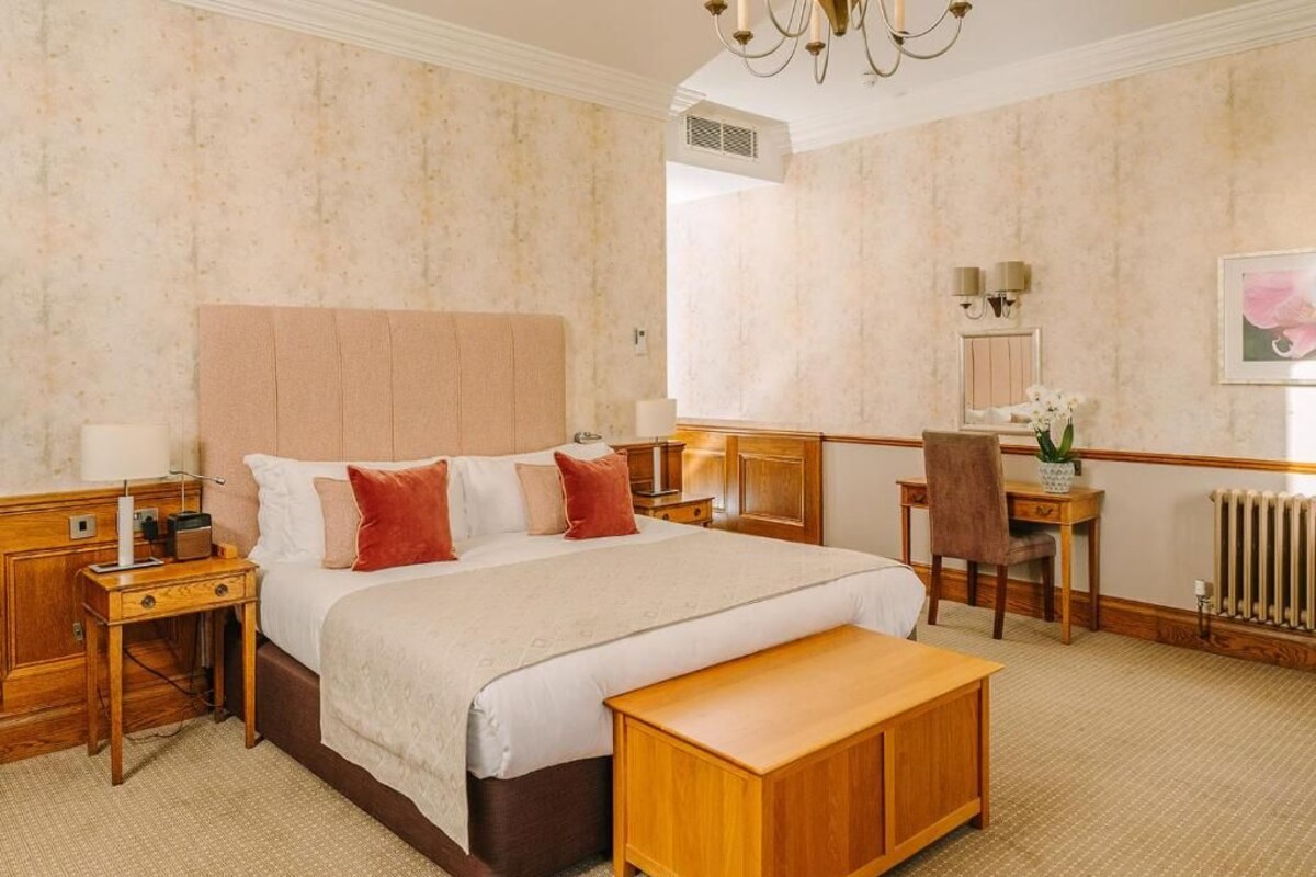 A hotel room with a large bed, bedside tables, a desk, a chair, a bench, and a radiator.